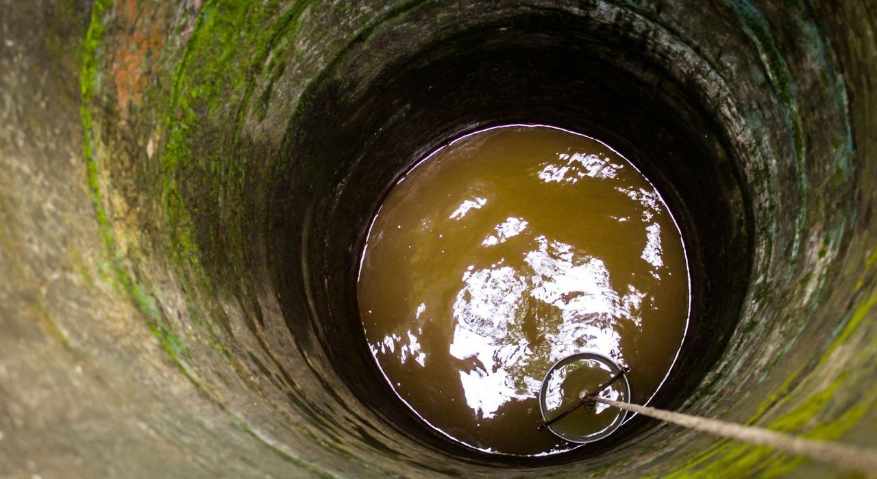 Interior de um antigo poço de pedra, com água turva, corda e balde.