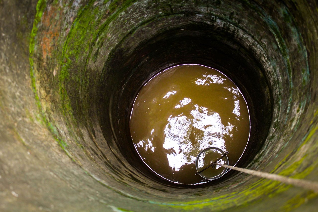 Interior de um antigo poço de pedra, com água turva, corda e balde.