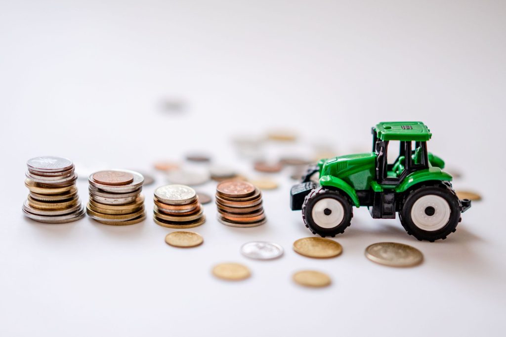 Miniatura de um trator verde ao lado de pilhas de moedas sobre uma superfície clara.