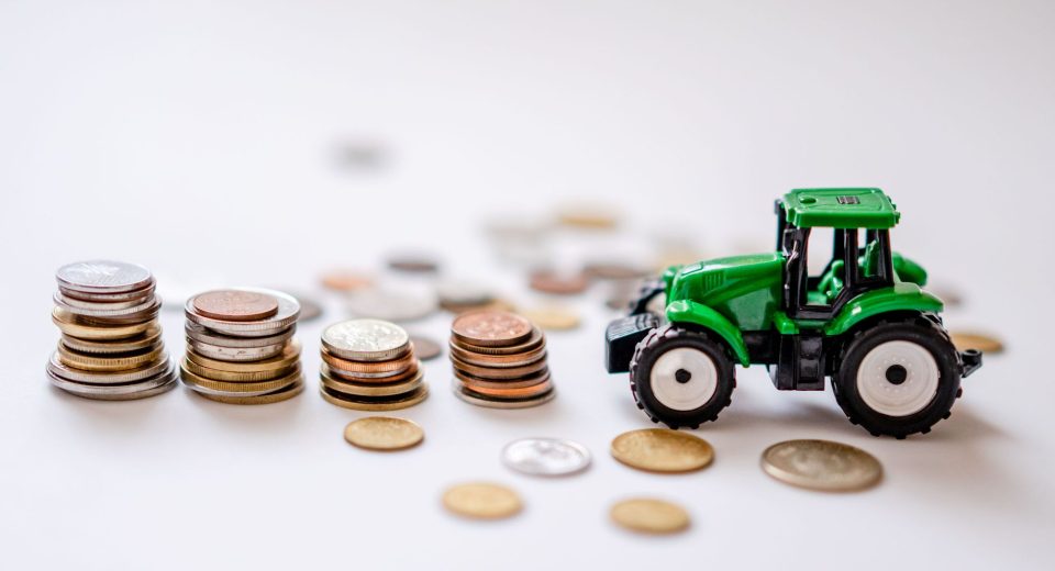 Miniatura de um trator verde ao lado de pilhas de moedas sobre uma superfície clara.